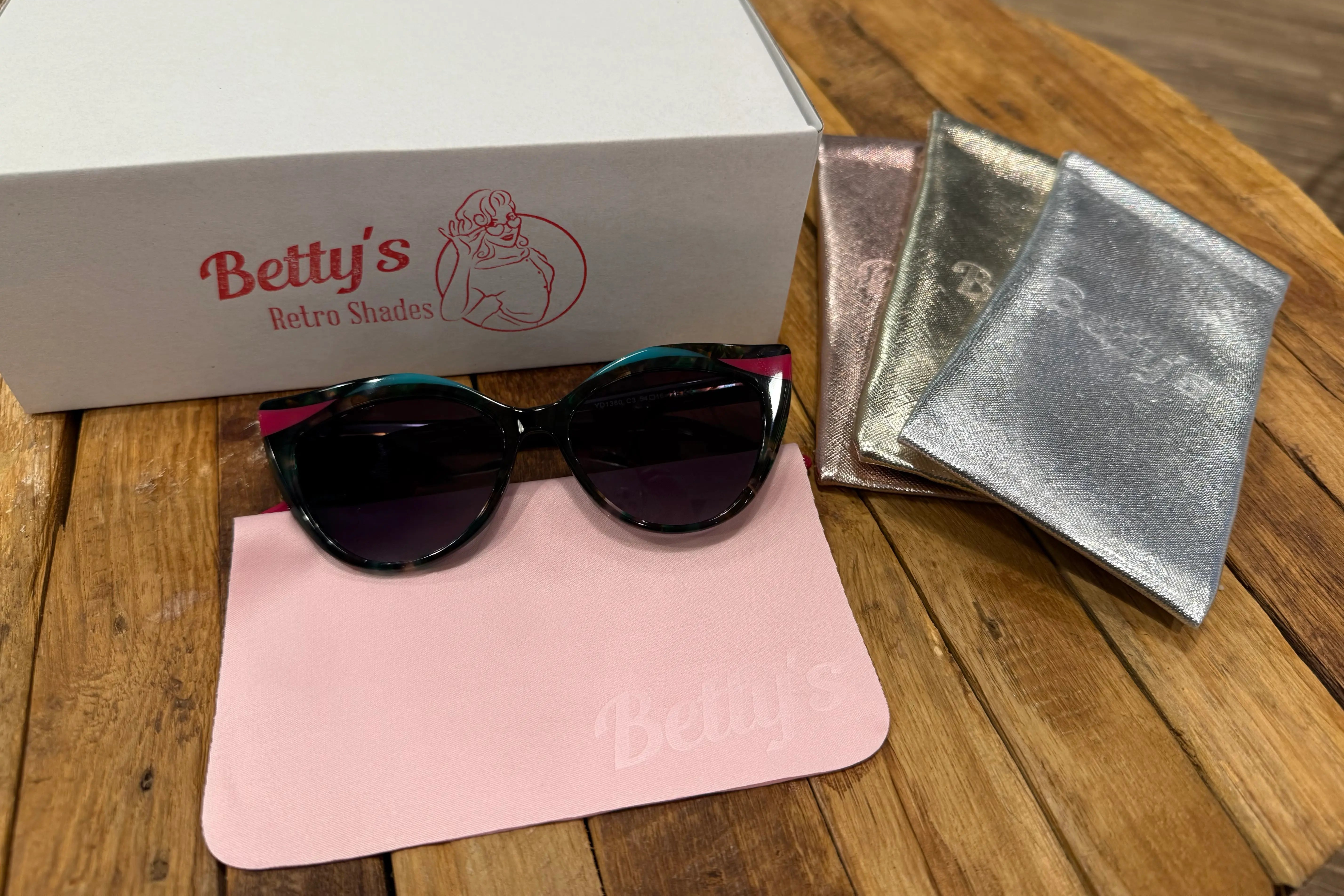 Sunglasses with a pink cloth and silver case on a wooden surface, branded 'Betty's'.